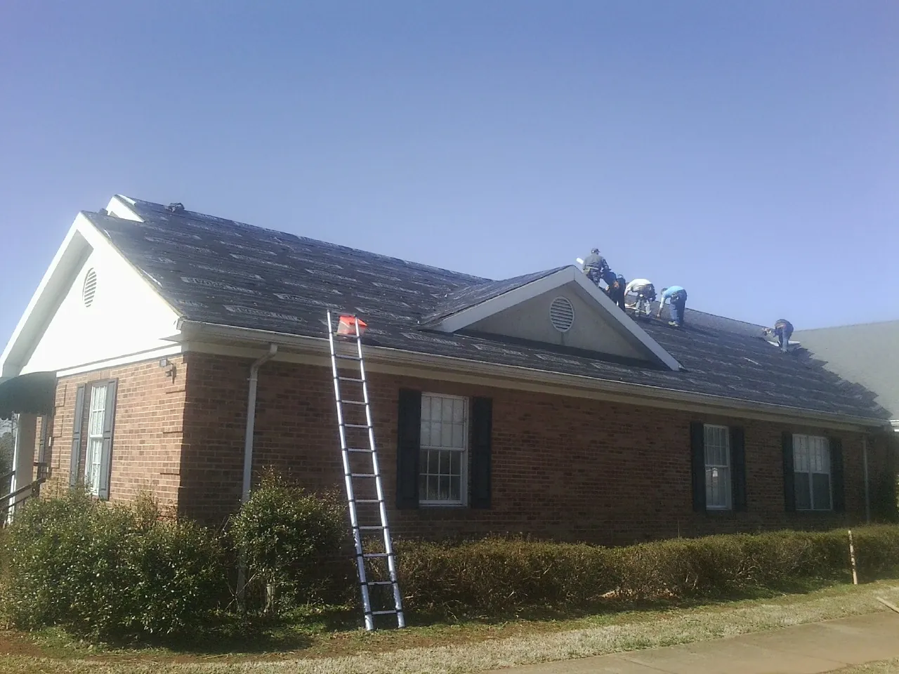 Crew tearing shingles off a brick ranch roof with a ladder set — Metal Roof Repair work by our crew in Candia