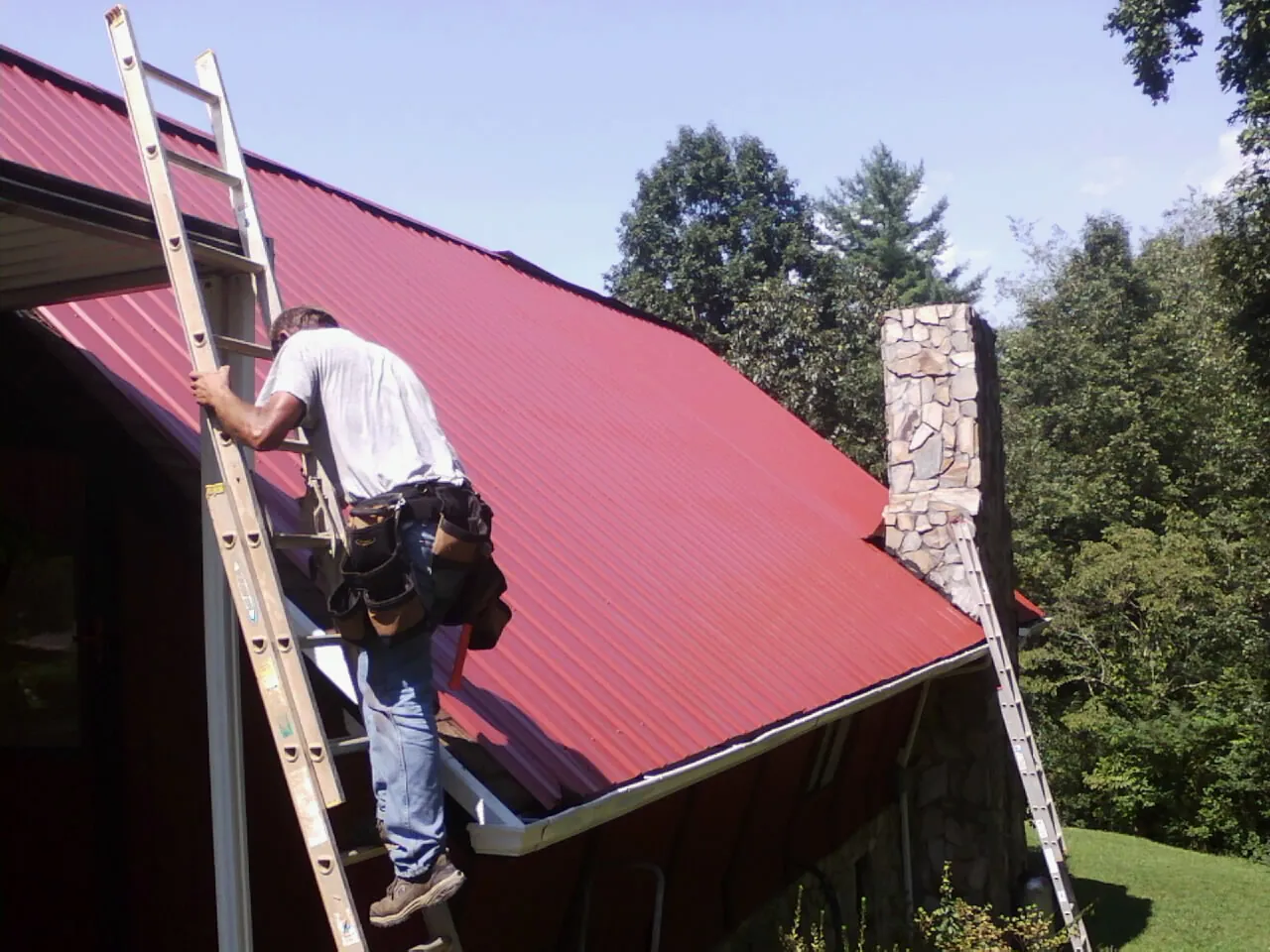 Roofer on a ladder beside a red metal roof and stone chimney — Soffit & Fascia Repair by Roofing Brothers in Candia